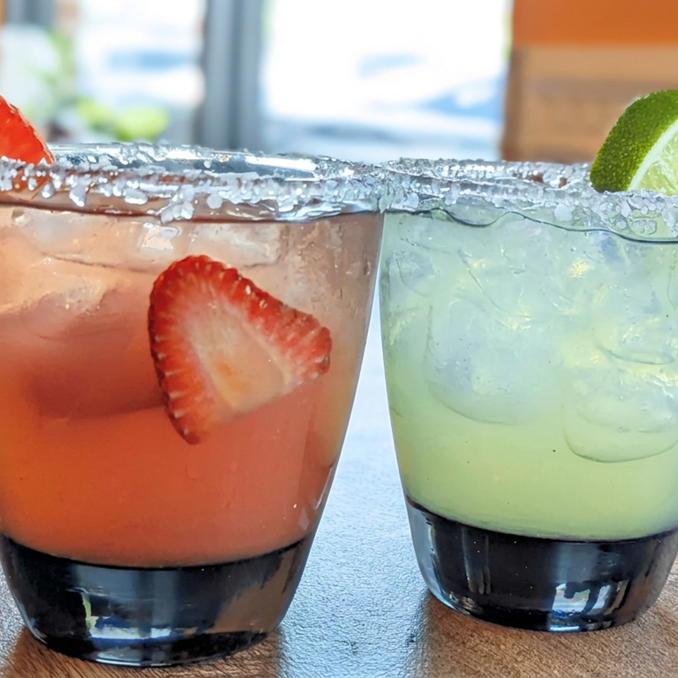 Two colorful iced cocktails with salted rims, garnished with strawberries and lime, sit beside a bowl of salsa on a wooden table.
