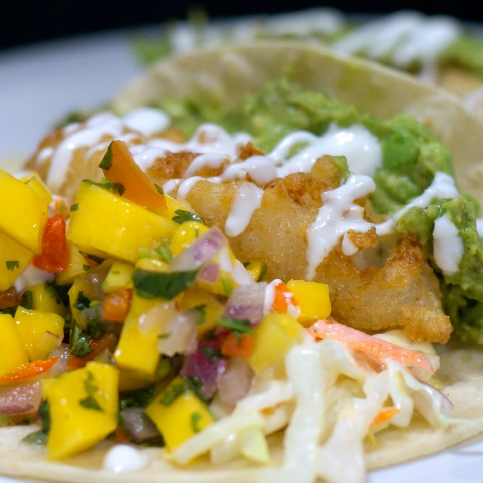 Two crispy fish tacos topped with lettuce, guacamole, creamy sauce, and vibrant mango salsa on a white plate.